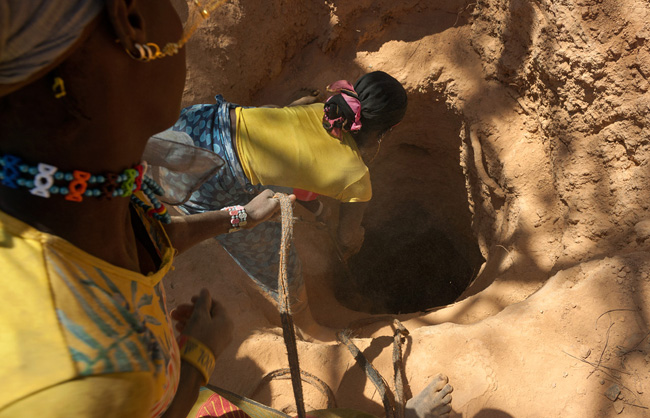 Two women are pulling out a basket full of stones from the above of 40 meters deep shaft. The first woman is on the edge of the shaft, fully stoop and legs wide apart. She takes this position because of the weight of the basket, to be able to pull it up. It is common that people lost their grips and fell countless meter down the shaft.