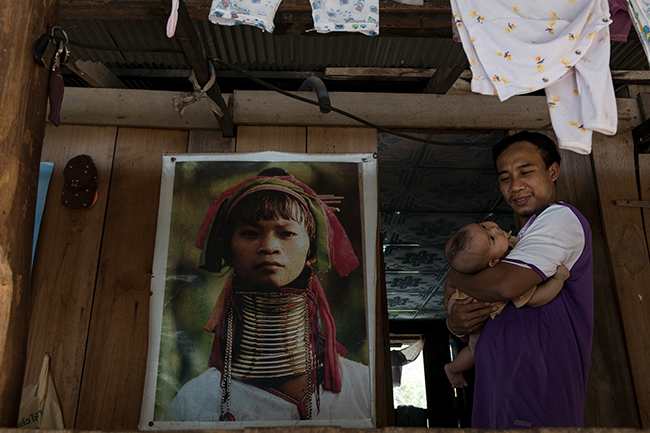 The images of the Kayan women and their metal ornaments around their necks have been seen around the world and have become icons of Northern Thailand tourism. Here, the teacher of the village’s only school, which was close due to Coronavirus restrictions, looks after his son in front of the iconic photo of his deceased mother.